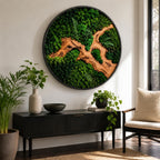 A circular wall art piece featuring a black frame filled with preserved green moss and a central piece of light brown driftwood. The moss displays various shades of green, including deep forest and bright lime, with small fern fronds.