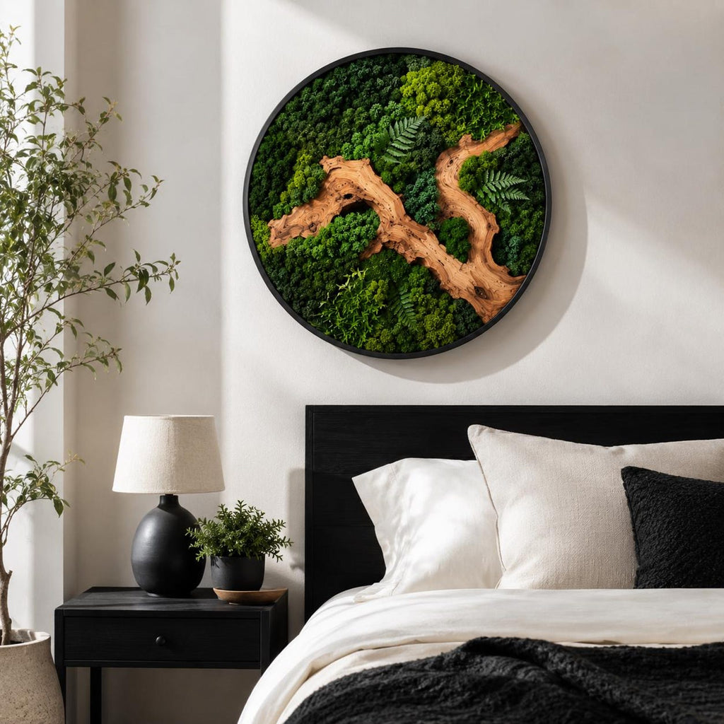 A circular wall art piece featuring a dense arrangement of preserved green moss in various shades, accented by a prominent, winding piece of light brown driftwood and delicate fern fronds, all enclosed within a thin black frame.