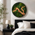 A circular wall art piece featuring a dense arrangement of preserved green moss in various shades, accented by a prominent, winding piece of light brown driftwood and delicate fern fronds, all enclosed within a thin black frame.