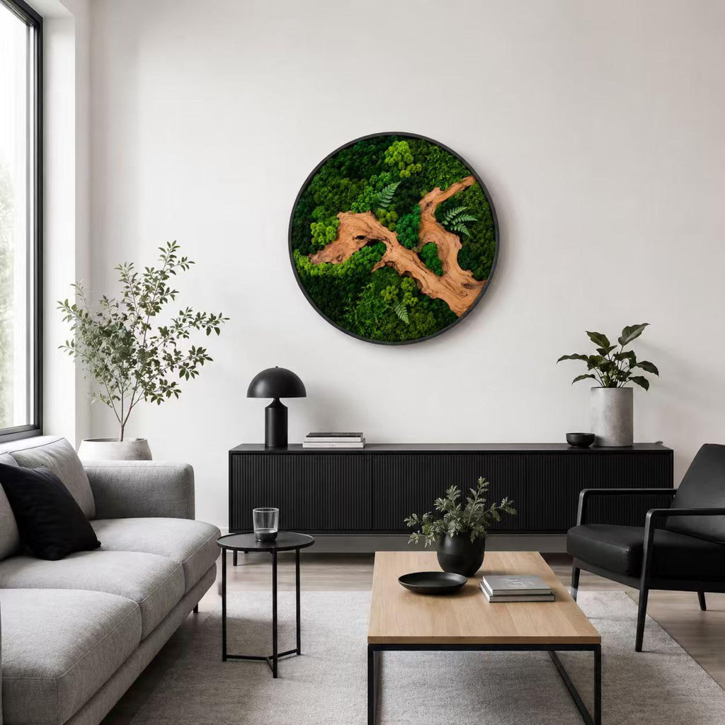 A circular wall art piece featuring preserved green moss and a central piece of light brown driftwood. The arrangement includes various shades of green moss and small fern-like leaves, all contained within a thin black circular frame.