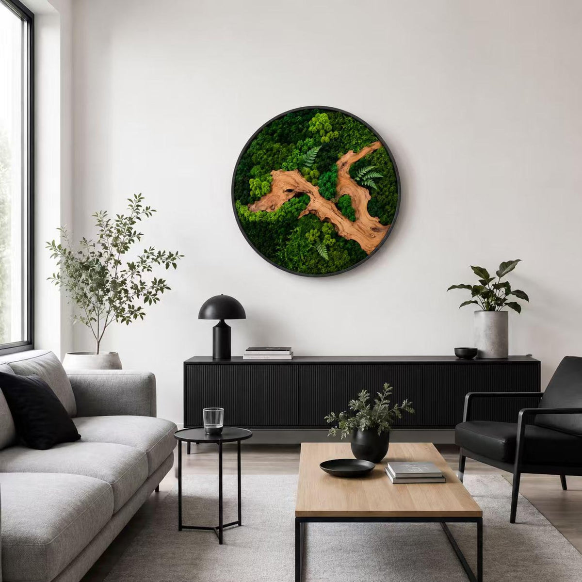 A circular wall art piece featuring preserved green moss and a central piece of light brown driftwood. The arrangement includes various shades of green moss and small fern-like leaves, all contained within a thin black circular frame.
