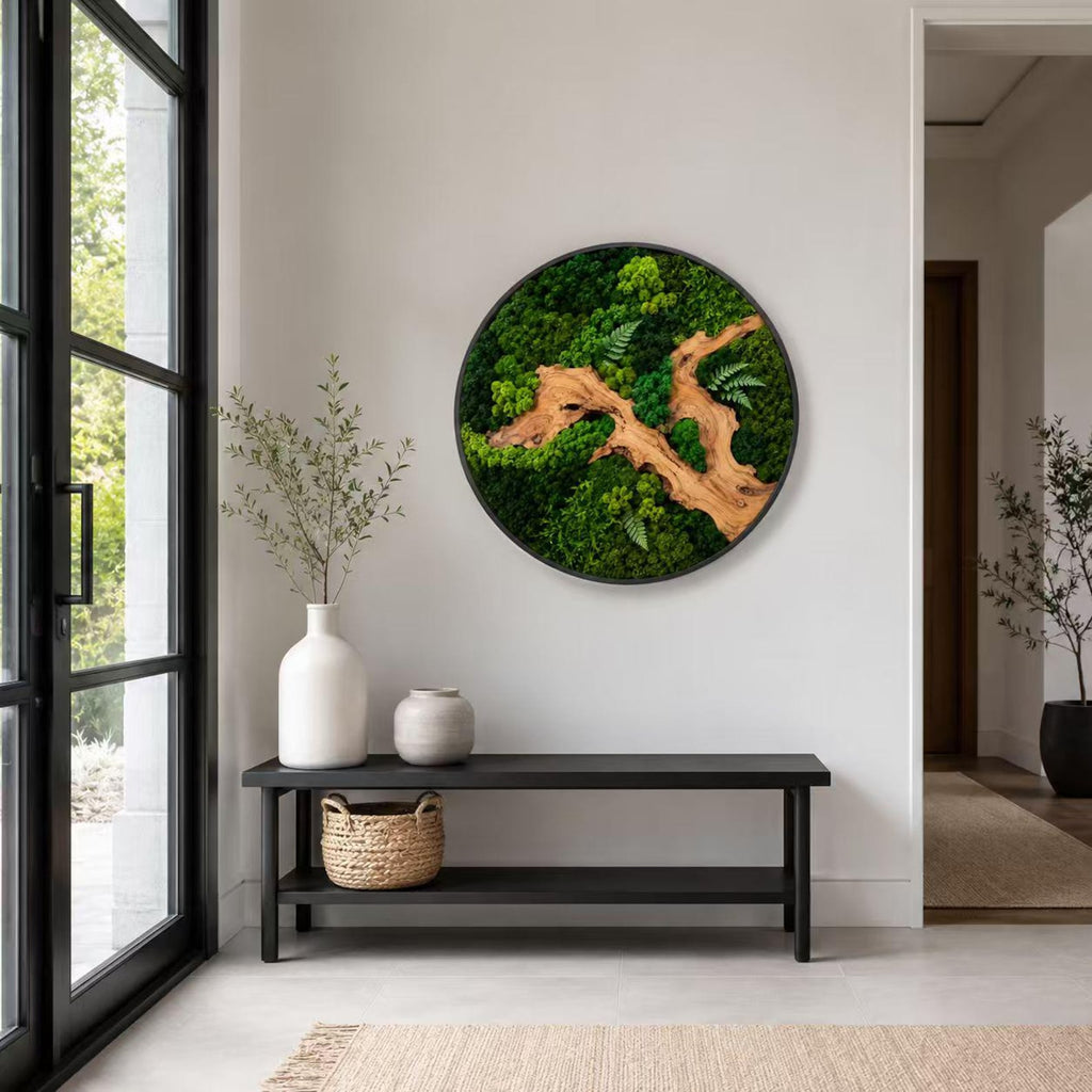 A circular wall art piece featuring preserved green moss and a central piece of light brown driftwood. The artwork is encased in a thin black frame and displays a textured, natural composition of various moss shades and fern leaves.