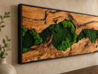 A rectangular wall art piece featuring a natural wood slab with a prominent grain pattern and dark fissures, inlaid with vibrant green preserved moss. The entire composition is encased in a thin, black metal frame.