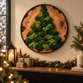 A circular wall art piece featuring a natural wood slice frame with a black metal border. The center displays a textured, three-dimensional Christmas tree shape crafted from preserved green moss in various shades of light and dark green.