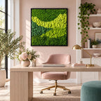 A square wall art piece featuring a dense arrangement of preserved moss in varying shades of vibrant lime green and deep forest green, encased in a thin black frame, mounted on a light pink wall above a wooden desk and chair.