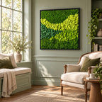 A square wall art piece featuring preserved moss in various shades of green, ranging from deep forest green to bright lime, arranged in a wavy, textured pattern within a thin, dark, rectangular frame mounted on a sage green wall.