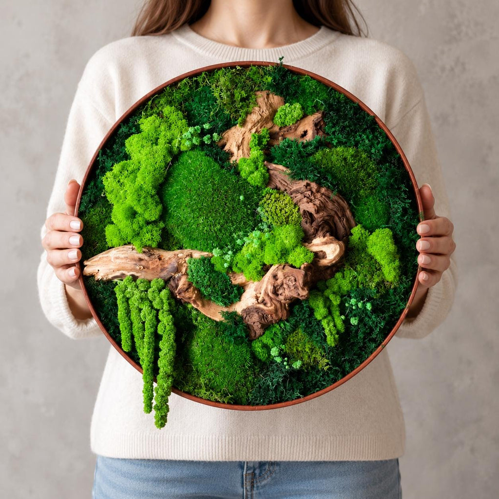 A circular wall art piece featuring a variety of preserved green mosses in different textures and shades, accented by pieces of natural brown driftwood. The arrangement is contained within a thin, round, dark-colored wooden frame.