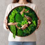 A circular wall art piece featuring a variety of preserved green mosses in different textures and shades, accented by pieces of natural brown driftwood. The arrangement is contained within a thin, round, dark-colored wooden frame.