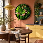 A circular wall art piece featuring preserved green moss and natural driftwood arranged in a dense, textured composition. The artwork is framed in a thin, dark circular border and mounted on a solid, warm-toned terracotta wall.
