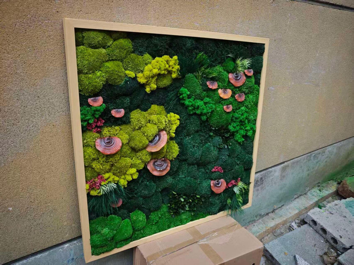 A square wall art piece featuring a variety of preserved moss in shades of lime, forest, and emerald green, accented with several small, circular, brown wooden mushroom-like decorative elements, all encased in a light-colored wooden frame.