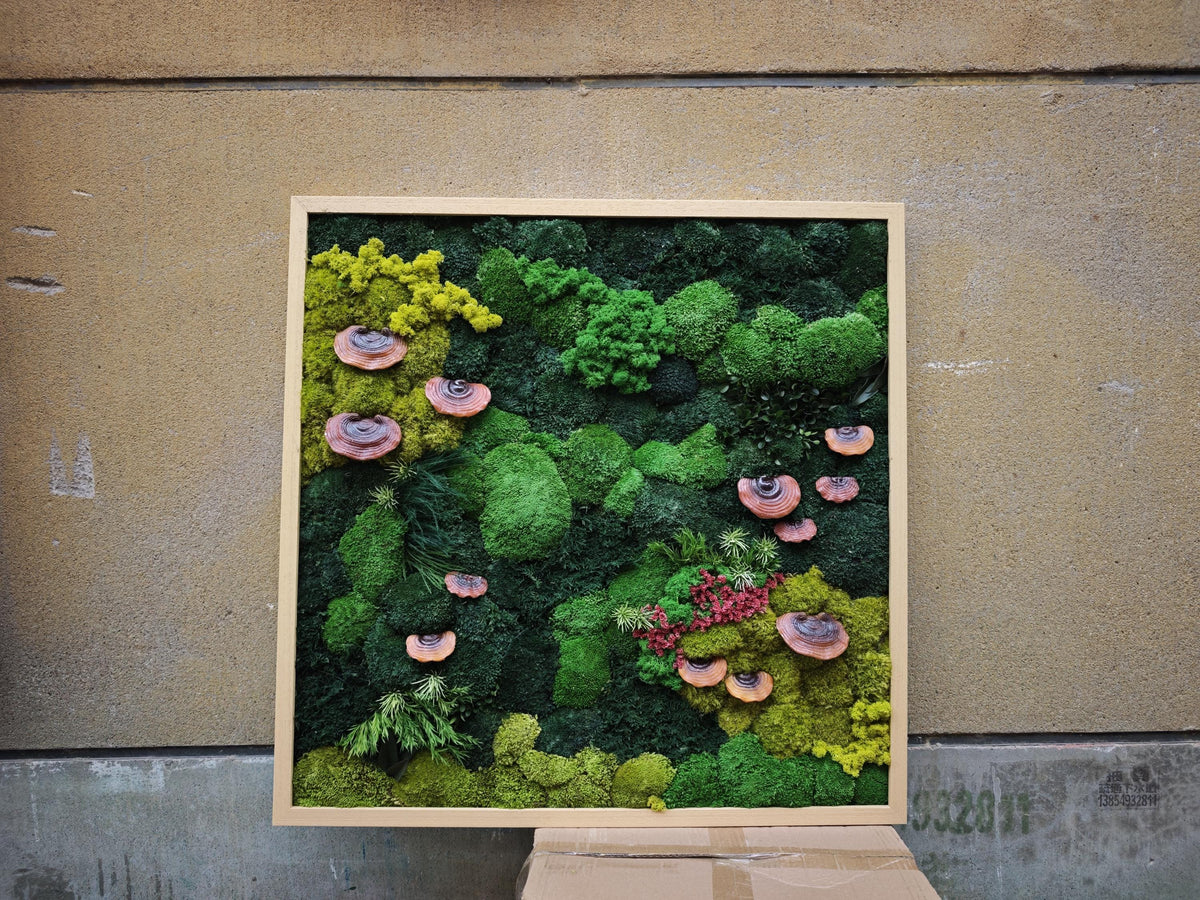 A square wall art piece featuring a wooden frame filled with preserved moss in various shades of green, ranging from lime to dark forest tones. Several brown, shelf-like fungi are attached throughout the textured moss arrangement.