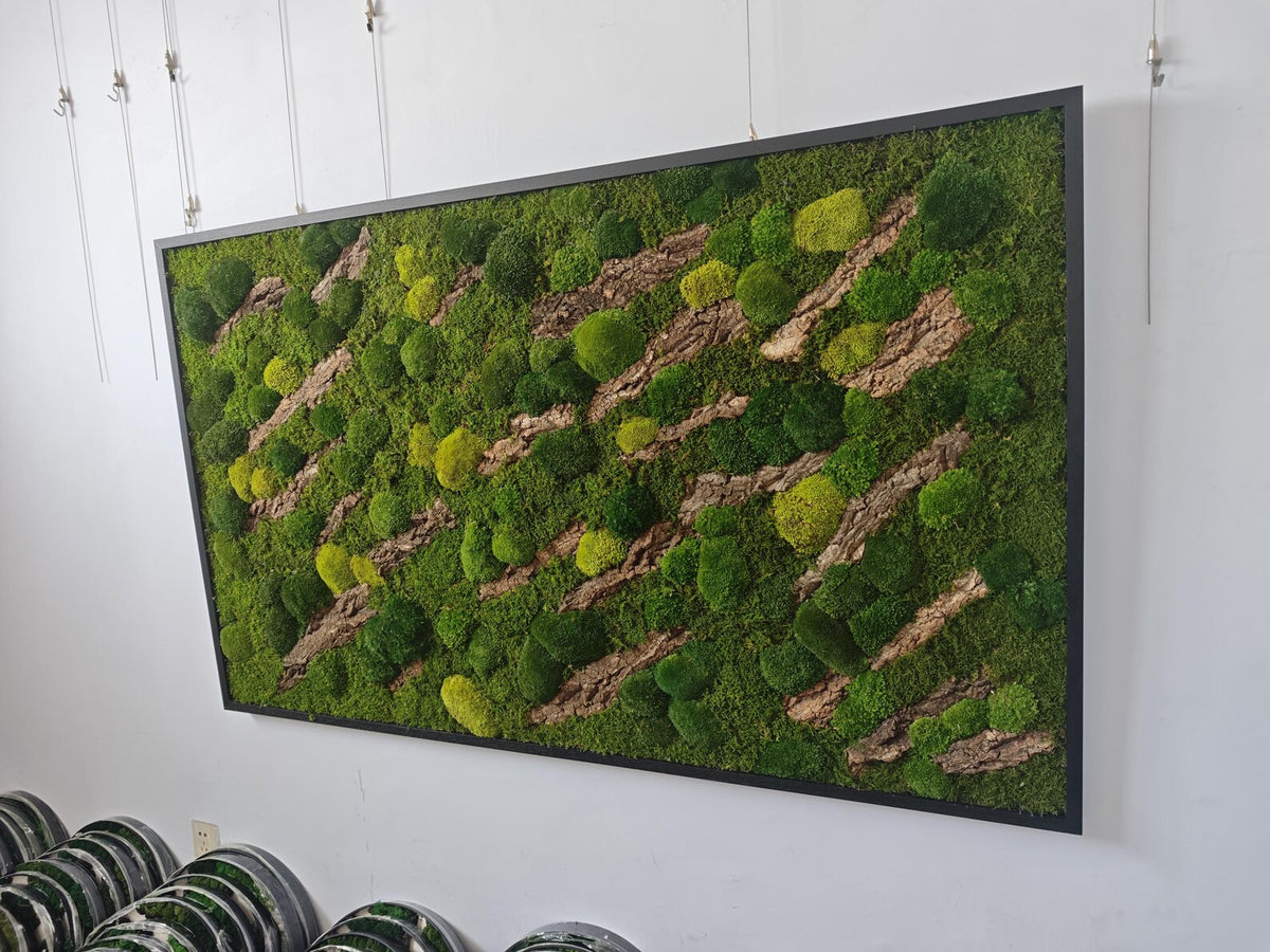 A rectangular wall art piece featuring a dense arrangement of preserved green moss in various shades, interspersed with diagonal strips of textured brown tree bark, all enclosed within a thin, black metal frame.