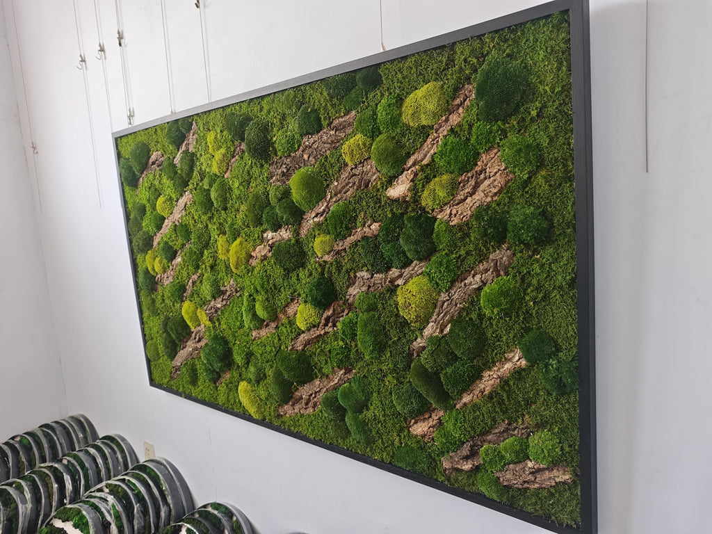 A rectangular wall art piece featuring a dense arrangement of preserved green moss in various shades, interspersed with diagonal strips of textured brown tree bark, all enclosed within a thin black metal frame.
