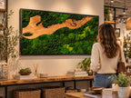 A rectangular wall art piece featuring a black frame enclosing a natural wood grain section surrounded by dense, vibrant green preserved moss. The composition creates a landscape effect with organic textures and varied shades of green.