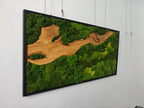 A rectangular wall art piece featuring a black frame containing a natural wood slab with a prominent grain pattern, surrounded by dense, textured green moss in varying shades of forest and lime green, creating a landscape effect.