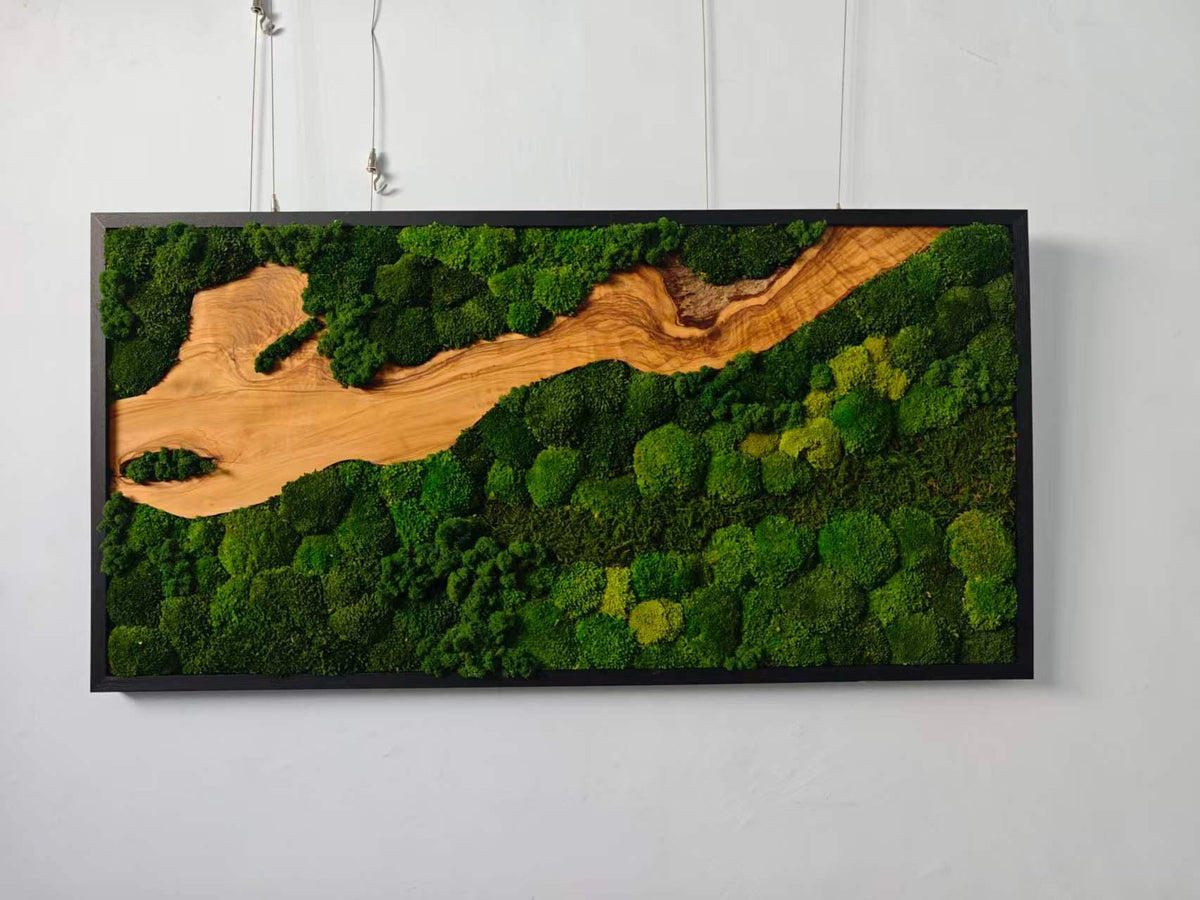 A rectangular wall art piece featuring a black frame enclosing a natural wood slab with a prominent grain pattern, surrounded by dense clusters of preserved green moss that create a textured, organic landscape across the surface.