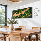 A rectangular wall art piece featuring preserved green moss and natural wood elements in a black frame. The wall displays the text "More Greenery A Kinder World." The artwork is positioned above a wooden dining table with a vase.